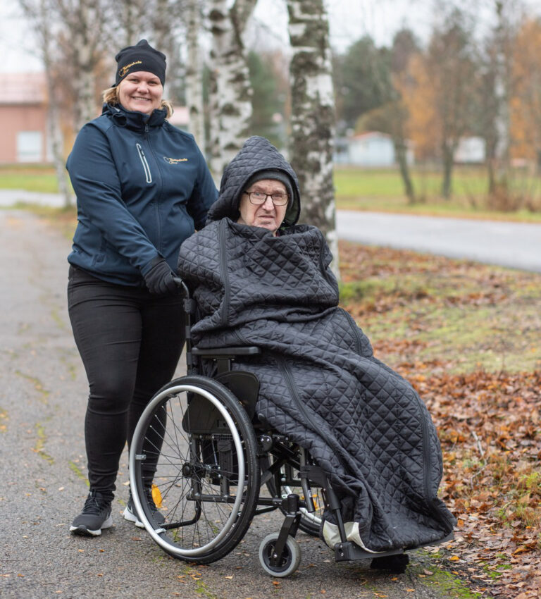 Kotipalvelu vanhuksille. Iloinen hoitaja seisoo ulkona pyörätuolissa istuvan vanhemman henkilön vieressä, molemmat katsovat kameraan. Pyörätuolissa oleva henkilö on peitetty lämpimällä huovalla, ja ympärillä on syksyinen maisema.