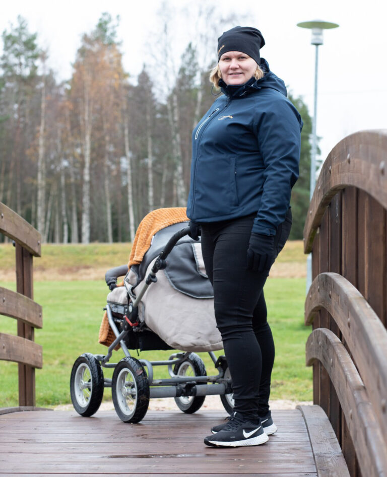 Kuvituskuva lapsiperheiden kotihoito. Nainen seisoo ulkona puusillalla lapsenrattaat vieressään. Hänellä on päällään tumma takki, mustat housut ja urheilukengät sekä päässään musta pipo. Taustalla näkyy vihreitä puita ja pilvinen taivas.